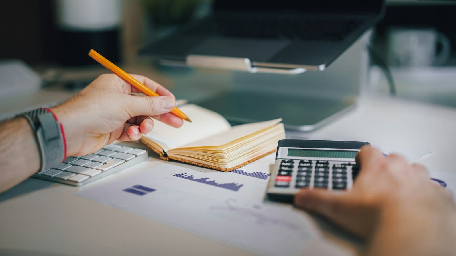 Mains tenant un crayon au-dessus d'un carnet, entre une calculatrice, des graphiques imprimés et un ordinateur portable