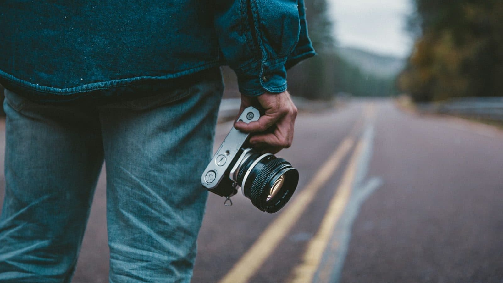 Photographe de dos tenant son appareil photo à la main, sur une route bordée d'arbres