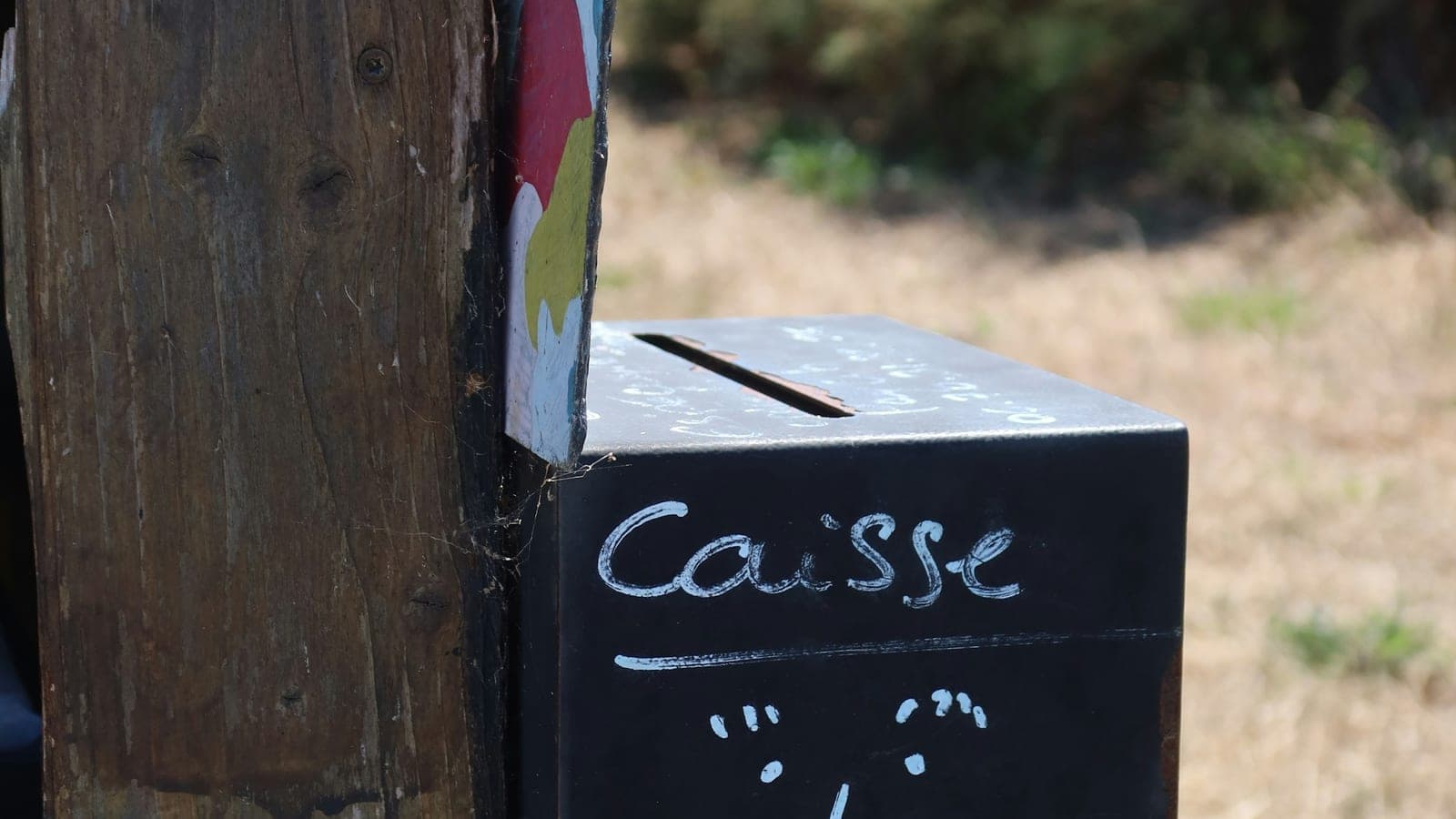 Boîte de collecte en métal noir fixée à un poteau en bois, avec le mot caisse écrit à la main