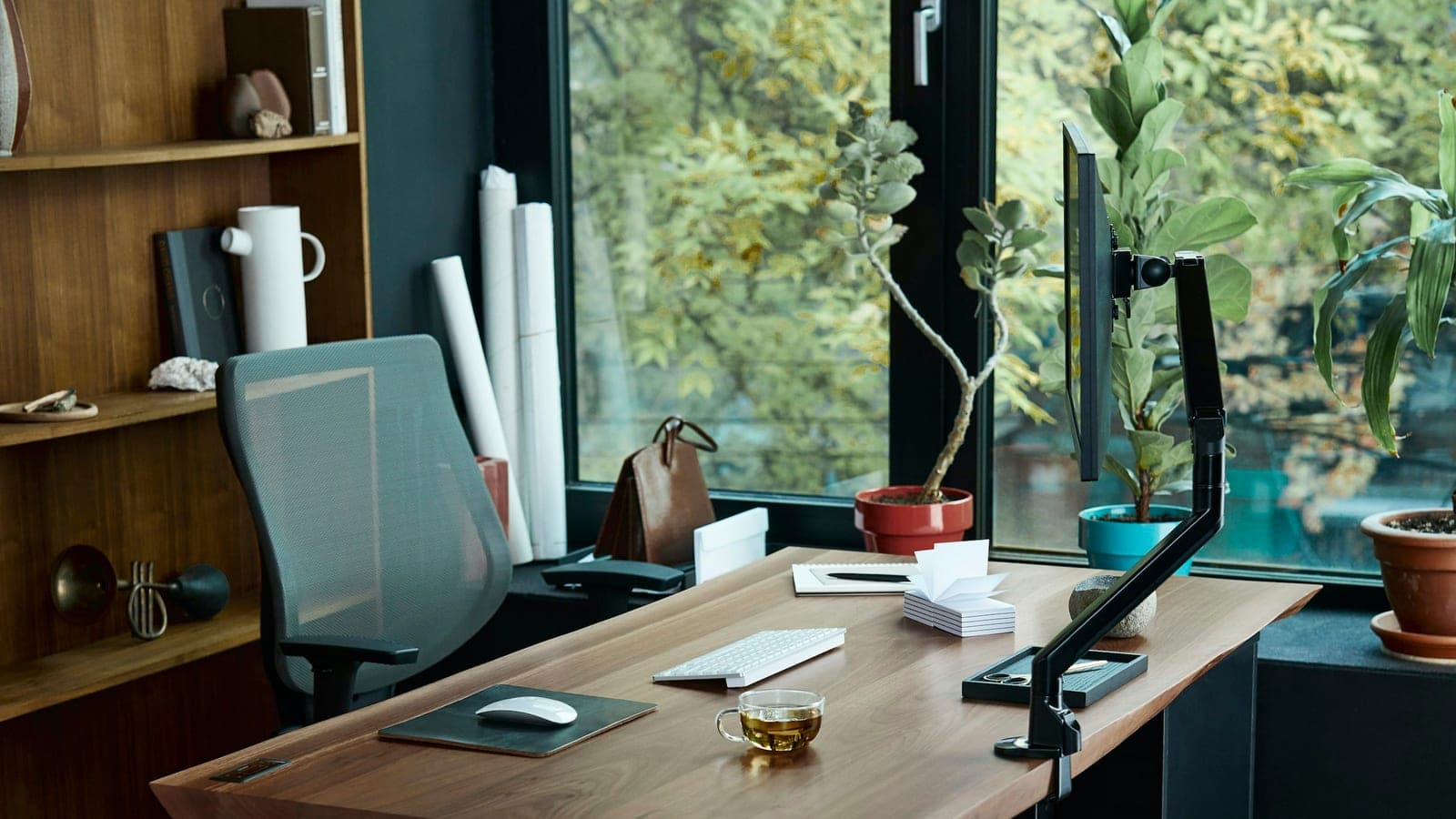 Bureau en bois clair devant une baie vitrée donnant sur des arbres, avec fauteuil de travail, écran sur bras articulé et plantes