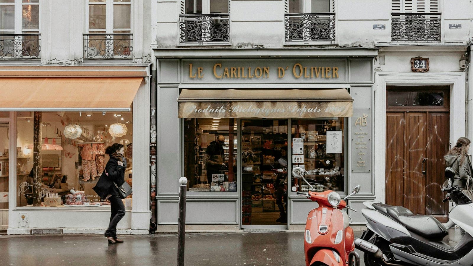 Devantures de commerces indépendants au rez-de-chaussée d'un immeuble parisien, sous la pluie, avec scooters garés le long du trottoir