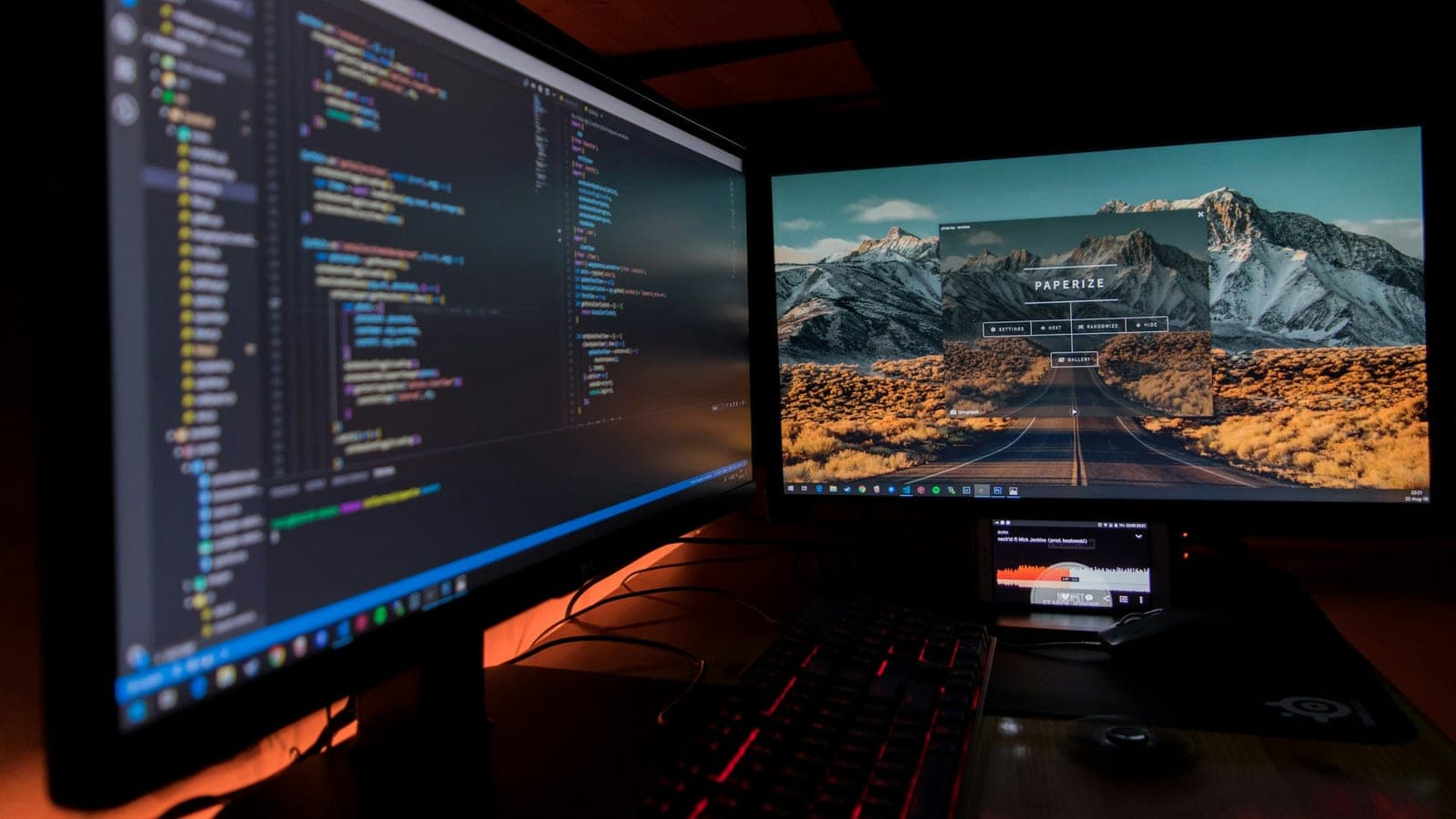 Deux écrans dont l'un affiche du code, sur un bureau, dans une pièce sombre