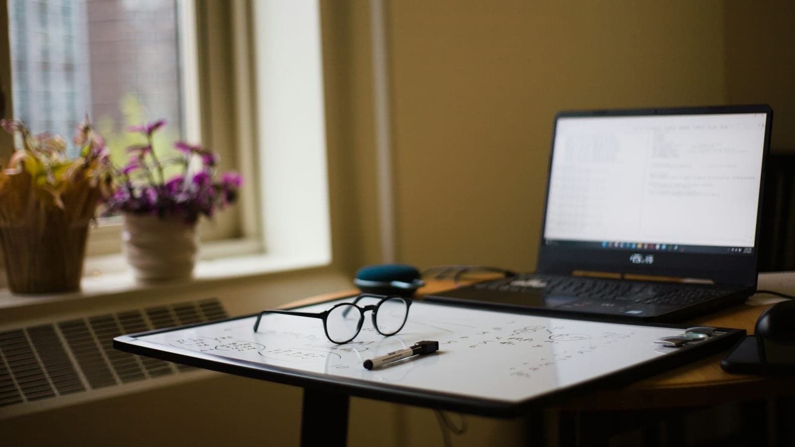 Ordinateur portable ouvert sur un bureau près d'une fenêtre, avec des lunettes et un feutre posés sur un tableau blanc