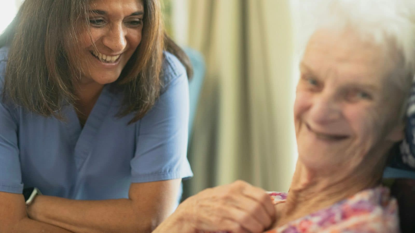 Infirmière en blouse bleue souriant au chevet d'une patiente âgée, à domicile