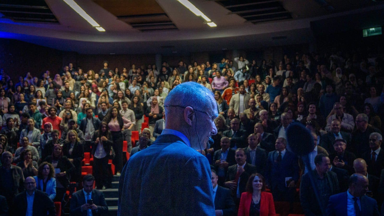 Intervenant de dos face à un amphithéâtre comble lors d'une conférence